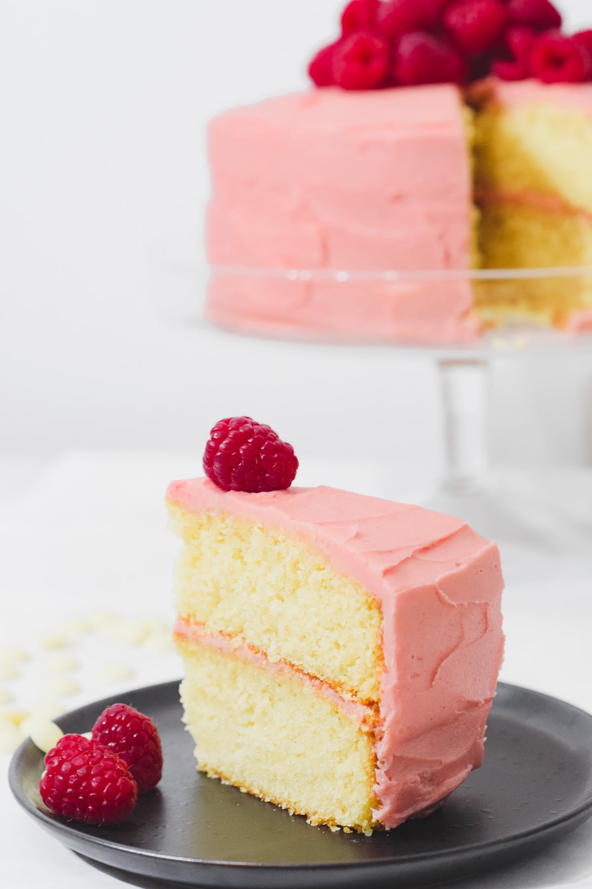 Slice of white chocolate cake with raspberry buttercream frosting a raspberry on top. Whole raspberry cake in the background.