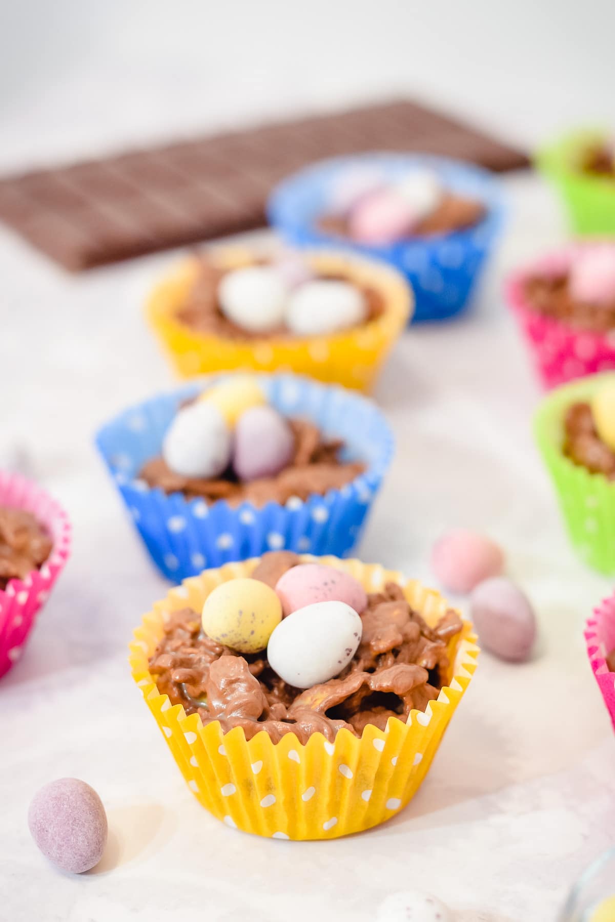Easter chocolate cornflake nest cakes in cupcake liners topped with mini eggs and chocolate in the background