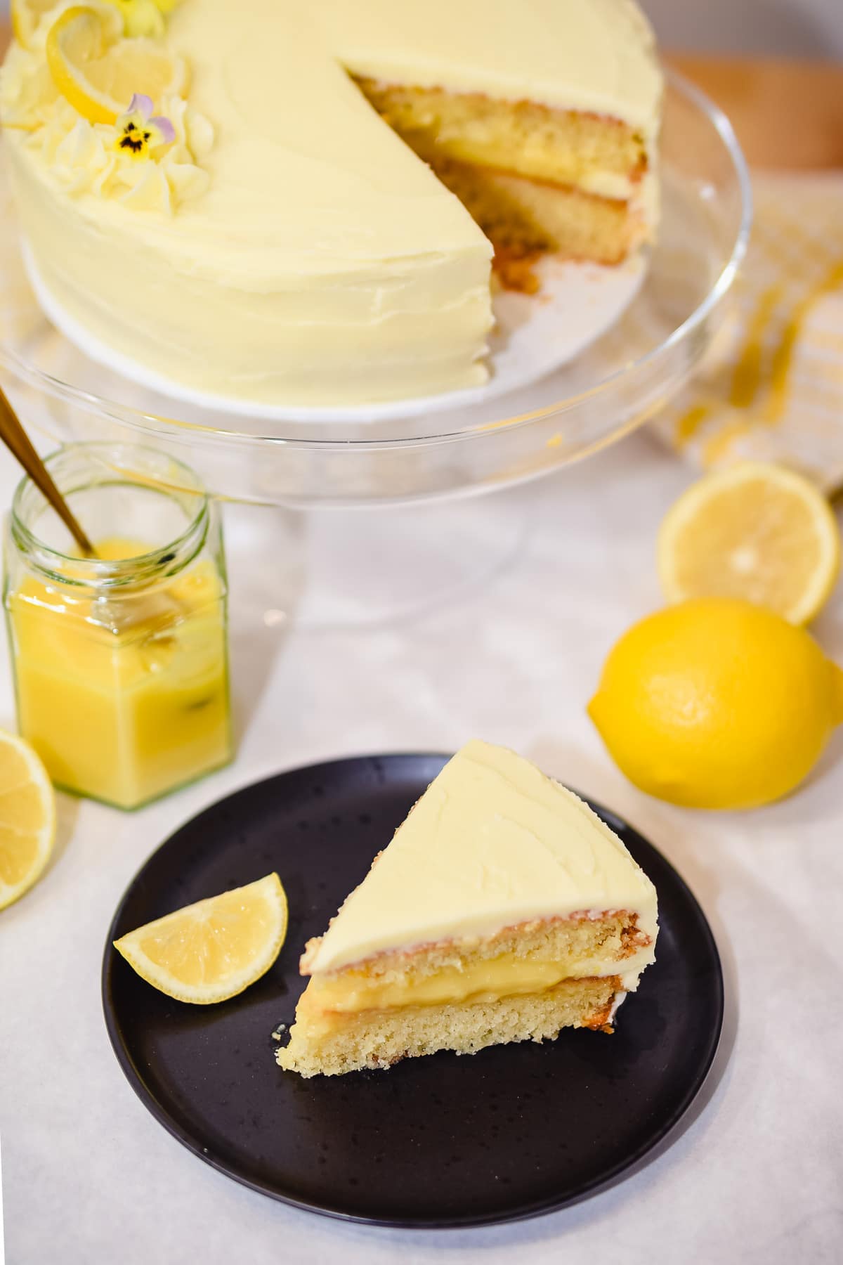 lemon curd cake slice on a plate with lemon cake on a cake stand behind
