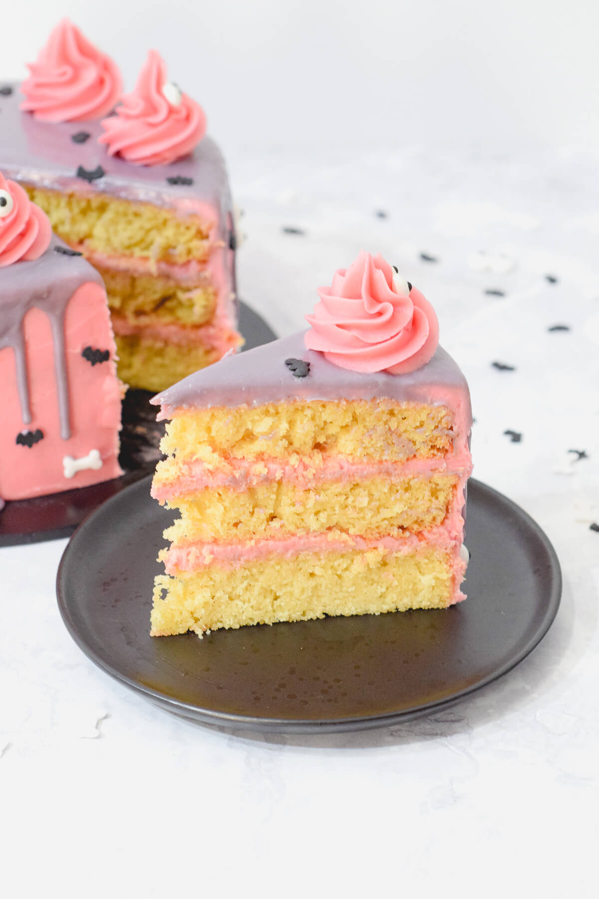 slice of pink halloween sponge cake showing three vanilla layers with a pink frosting and filling with pink monsters on top.