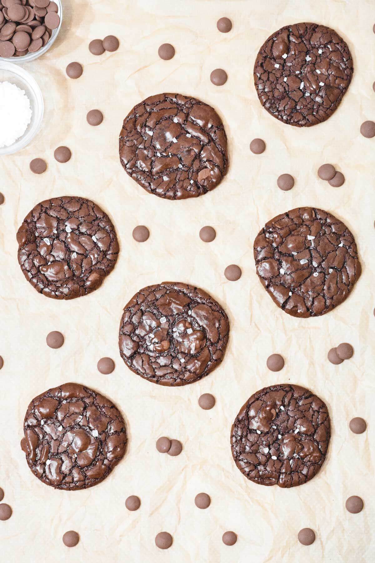gluten free chocolate brownie cookies with chocolate chips and sprinkled with sea salt with bowls of salt and chocolate behind.